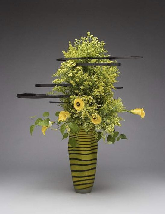 A floral arrangement that uses a striped vase. Yellow flowers and green foliage come up from the base, while wooden bars are staggered horizontally across the foliage